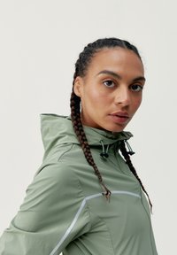 Svetlozelená, ľahká bunda s kapucňou; obsahuje reflexné akcenty a sťahovaciu šnúrku. Hladká textúra, prispôsobený strih.
