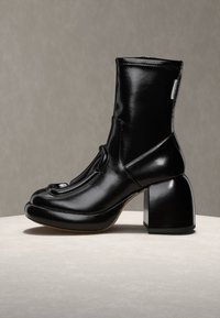 Botte en cuir noir brillant mi-mollet avec un talon carré bloc et un bout arrondi, posée sur une surface claire contre un fond neutre.