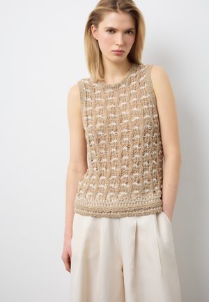 Jeune femme aux cheveux blonds portant un haut tricoté sans manches beige et un pantalon large écru, debout devant un fond blanc.