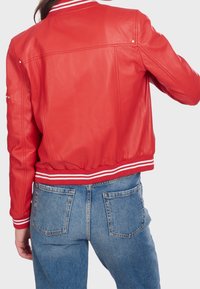 Osoba ubrana w czerwoną skórzaną kurtkę varsity z białymi paskami i niebieskie dżinsy, pokazana z tyłu na tle jednolitym.