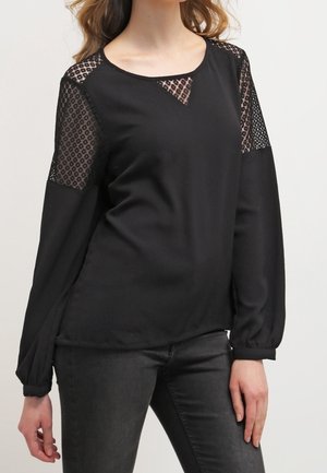 Woman wearing a black long-sleeve top with sheer patterned panels on shoulders and upper chest, paired with black jeans.