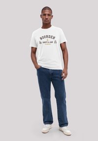 Weißes Baumwoll-T-Shirt mit dunkelbraunem Drucktext "NOORDEN," kombiniert mit blauen Jeans und weißenSneakers. Glatte Textur auf der gesamten Fläche.