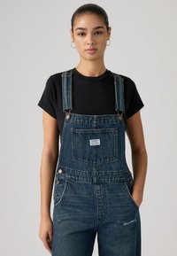 Νεαρή γυναίκα με σκούρα μαλλιά σε κότσο, που φορά χρυσούς κρίκους σκουλαρίκια, μαύρο μπλουζάκι και μπλε τζιν σαλοπέτα με τα χέρια στις τσέπες, ουδέτερο φόντο.