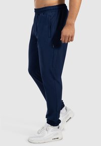 Tmavě modré sportovní kalhoty s elastickým pasem, bočními kapsami a úzkými kotníky, spárované s bílými sportovními botami. Hladká textilka.