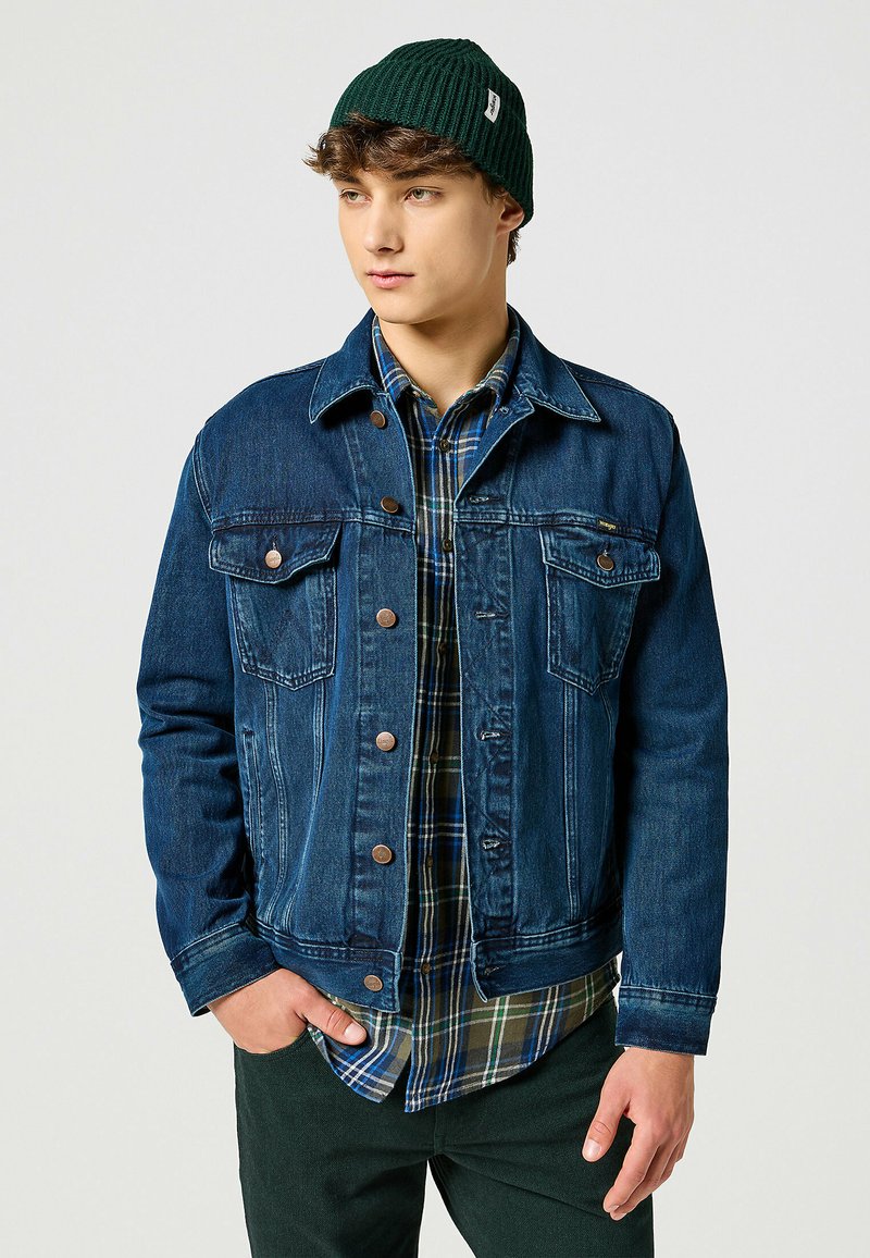 Chaqueta de mezclilla azul oscura con dos bolsillos en el pecho, botones de metal y un collar estructurado. Llevada sobre una camisa de cuadros verdes y pantalones oscuros.