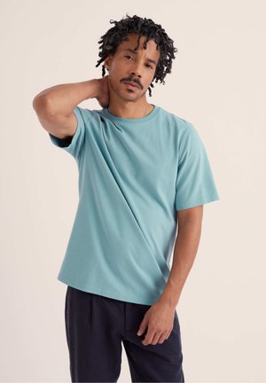 Göndör hajú férfi világoskék pólóban és sötét nadrágban, egyik kezével a nyaka mögött áll, semleges arckifejezéssel.