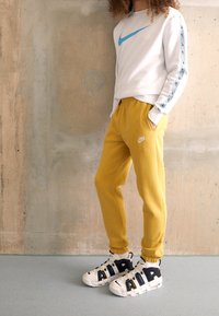 Nike långärmad tröja i vitt med blå logotyp, kombinerad med gula sweatpants med logotyp och vita sneakers med svarta detaljer.