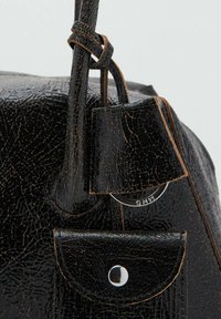 Primer plano de un bolso de mano de cuero negro agrietado con correa y un pequeño bolsillo con solapa que presenta un botón plateado y un detalle de anillo de metal.