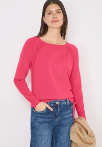 Femme aux cheveux longs et foncés portant un haut rose vif à manches longues et un jean bleu, tenant une veste beige dans sa main gauche.