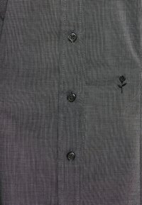Camicia grigia a bottoni con un sottile motivo a scacchi, dotata di bottoni neri e un piccolo ricamo di rosa nera vicino al petto.