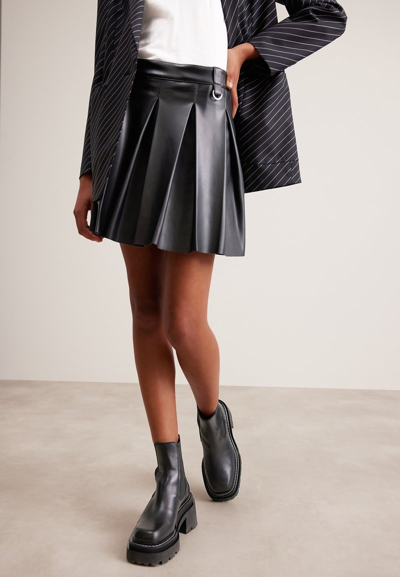 Personne portant une jupe plissée en cuir noir, des bottines noires épaisses, un haut blanc et un blazer noir à fines rayures avec la main sur la hanche.