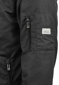 Musta nailonitakki, jossa on vetoketjullinen tasku tarralla, sileä pinta ja muotoiltu malli nykyaikaista ilmettä varten.