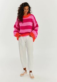 Oversized pulovr s růžovými a fuchsiovými pruhy, žebrovanými oranžovými manžetami a širokým výstřihem. Stylizováno s bílými kalhotami a béžovými teniskami.