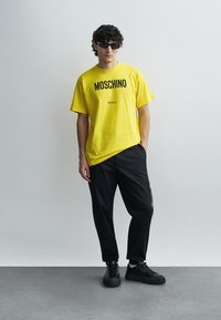 Žlté bavlnené tričko s čiernym nápisom „MOSCHINO Milano“, kombinované s čiernymi voľnými nohavicami a čiernymi teniskami. Doplnok: tmavé slnečné okuliare.