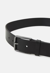 Svart läderbälte med en slät yta, försett med en silvermetallspänne och flera justeringshål längs hela sin längd.