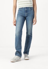 Ανοιχτό γαλάζιο τζιν παντελόνι με ίσια γραμμή και φθαρμένη εμφάνιση, πέντε τσέπες και κλασική μέση. Συνδυασμένο με γκρίζα αθλητικά παπούτσια.