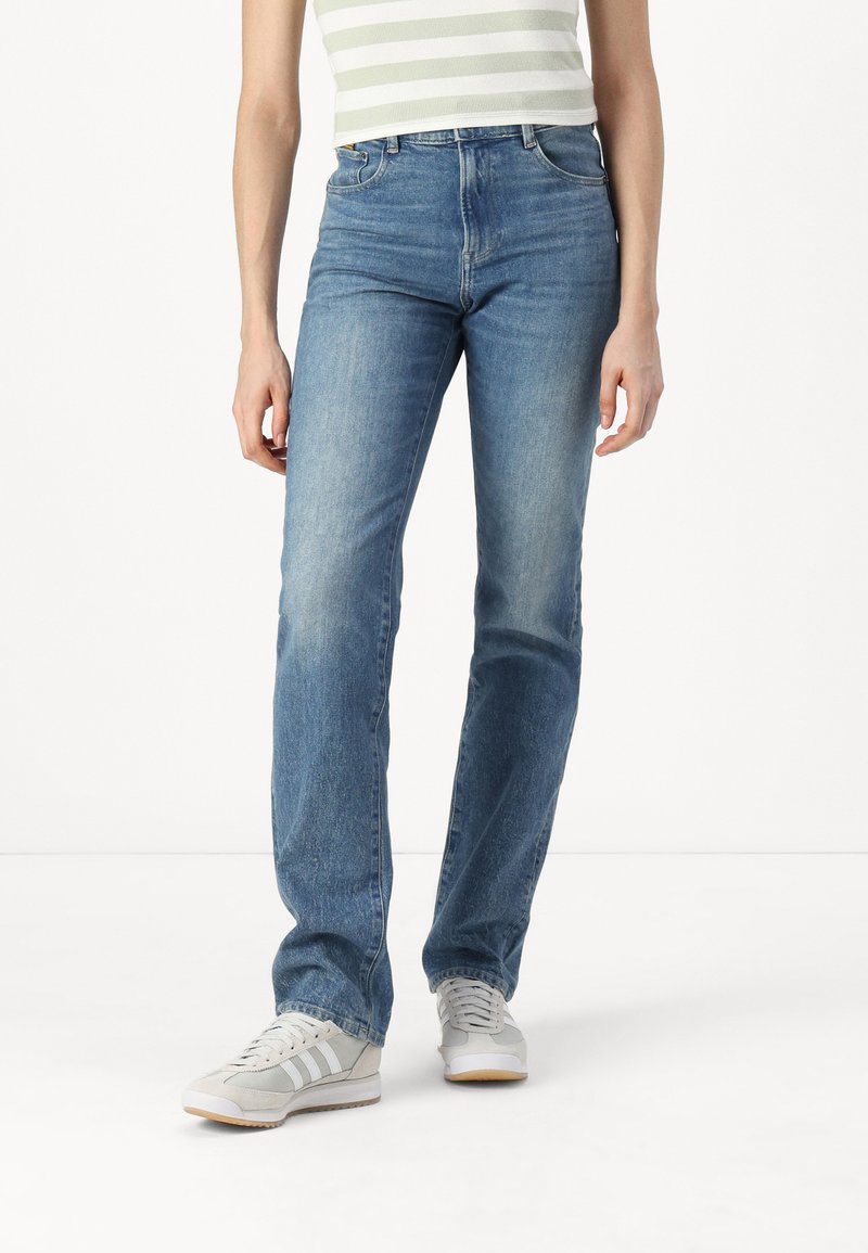 Ανοιχτό γαλάζιο τζιν παντελόνι με ίσια γραμμή και φθαρμένη εμφάνιση, πέντε τσέπες και κλασική μέση. Συνδυασμένο με γκρίζα αθλητικά παπούτσια.