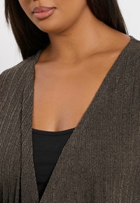 Brun ribbad cardigan med en djup V-ringning, lagd över en svart linne, som visar upp en strukturerad tyg.