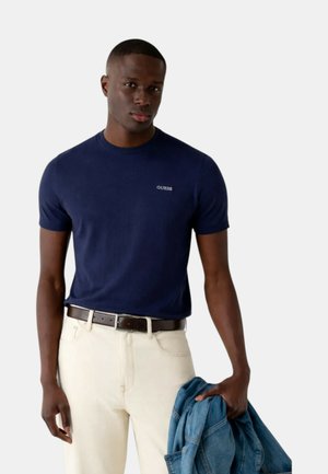 Man draagt een marineblauw Guess T-shirt en lichtgekleurde broek, houdt in zijn linkerhand een spijkerjasje vast tegen een witte achtergrond.