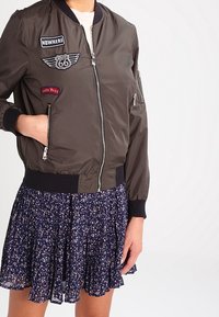 Giacca bomber verde oliva con toppe, chiusura con zip e polsini e orlo a coste, indossata sopra una gonna floreale blu navy.