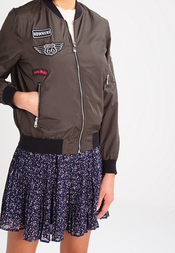 Giacca bomber verde oliva con toppe, chiusura con zip e polsini e orlo a coste, indossata sopra una gonna floreale blu navy.