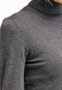 Zbliżenie na osobę noszącą dopasowany, ciemnoszary, prążkowany sweter z golfem i długimi rękawami.