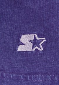 Violet medžiaga su siuvinėtu logotipu, kuriame yra stilizuota raidė „S“ ir žvaigždutė šviesiai violetinėje spalvoje. Glotni tekstūra, arti detalė.