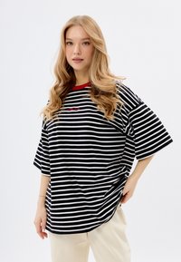 Czarny i biały paskowany, oversize'owy t-shirt z czerwonym akcentem przy kołnierzyku, z logo na przodzie i krótkimi rękawami.