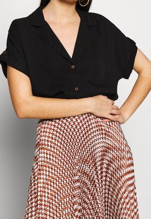 Kobieta nosząca złote kolczyki koła, czarną koszulę na guziki z krótkim rękawem i plisowaną spódnicę w brązowo-biały wzór pepitki, z założonymi na wysokości talii skrzyżowanymi ramionami.