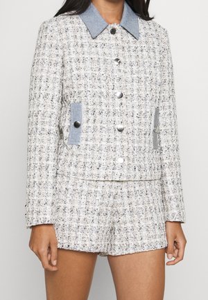 Frau trägt eine strukturierte weiße und schwarze Tweedjacke und passende Shorts mit silbernen Knöpfen und hellblauen Stoffakzenten an Kragen und Taschen.