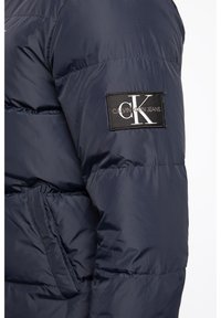 Marinblå puffjacka med en svart rektangulär lapp med vit "Calvin Klein Jeans" logotyp och en framträdande "CK" emblem på ärmen.