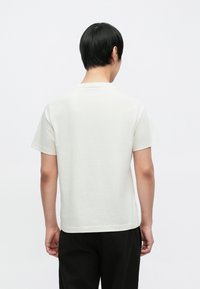 Tričko s krátkými rukávy a kulatým výstřihem v režném odstínu z bavlny, s volnějším střihem a rovným lemem. Pohled zezadu ukazuje minimální detaily.