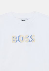 Λευκό βαμβακερό μπλουζάκι με τη λέξη "BOSS" σε βαθμιδωτό χρώμα από ανοιχτό μπλε έως μπεζ. Στρογγυλή λαιμόκοψη, κοντά μανίκια, λεία υφή.