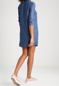 Kvinna iklädd en blå denimklänning med trekvartsärmar och ljusrosa sneakers, stående med ett ben något lyft.