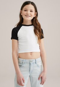 Top corto blanco acanalado con mangas cortas negras, combinado con unos vaqueros de cintura alta de color azul claro. El conjunto presenta un diseño simple y un ajuste cómodo.