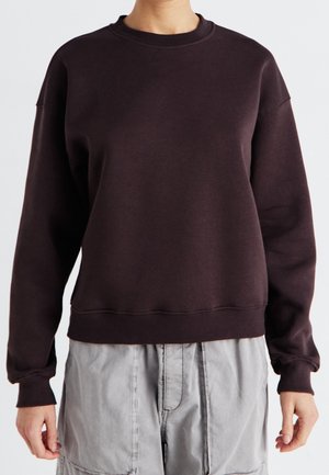 Osoba ubrana w ciemnobrązową bluzę z długim rękawem i okrągłym dekoltem oraz jasnoszare spodnie, pokazana od ramion do bioder na jednolitym tle.