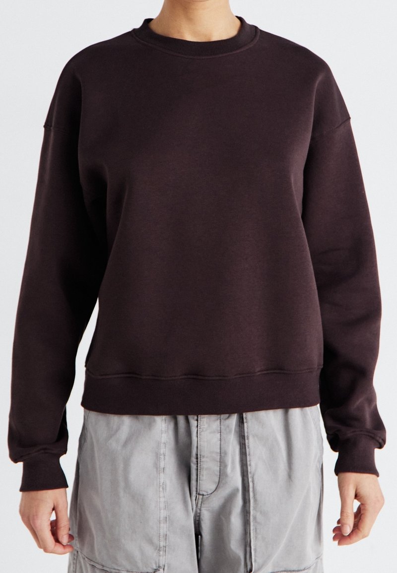 Personne portant un sweat-shirt à manches longues et col ras du cou de couleur brun foncé et un pantalon gris clair, visible des épaules aux hanches sur un fond uni.