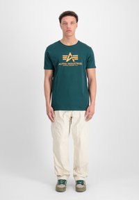Teal bomuld t-shirt med gyldent "ALPHA INDUSTRIES" logo, parret med cremede cargo bukser og grønne sneakers med netdetaljer.