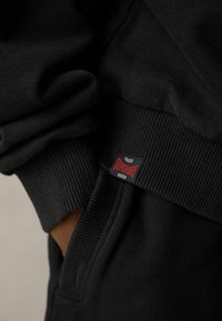 Svart sweatshirt med ribbstickade muddar och midja. En Superdry-etikett med röd text syns nära sömmen. Mjuk, strukturerad tyg.
