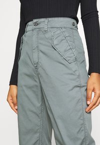 Pantaloni cargo grigi chiari con vita alta, tasche laterali e una texture liscia, leggermente stropicciata. Presenta chiusura con bottone e zip frontale.