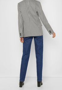 Persoon in een zwart-wit houndstooth blazer, blauwe jeans en zwarte enkelhoge hakken, staand met de rug naar de camera op een effen achtergrond.