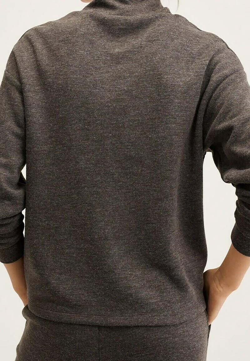 Personne portant un pull à manches longues gris foncé avec un col montant et un pantalon assorti, mains posées sur les hanches, de dos.