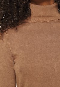 Brauner, gerippter Rollkragenpullover, der eine enge Passform mit hohem Kragen betont. Der Stoff erscheint weich mit einem leichten Glanz und minimalen Nähten.