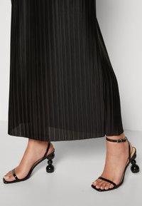 Černá plisovaná sukně spojená s černými sandály na řemínky s kulatým podpatkem zdobeným korálky. Hladká textura a čtvercový tvar špičky.