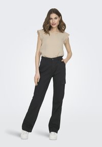 Haut beige sans manches avec dentelle, associé à un pantalon cargo noir avec poches latérales et des baskets blanches. Fond uni.