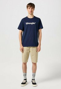 Marineblå bomulds T-shirt med "Wrangler" logo, sammen med beige shorts, grå sokker og sorte løbesko. Enkel, afslappet påklædning.