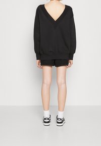 Zwarte sweatshirt met een diepe V-achterkant, ribgebreide manchetten en zoom, gecombineerd met zwarte shorts. Model draagt zwart-witte sneakers en sokken.