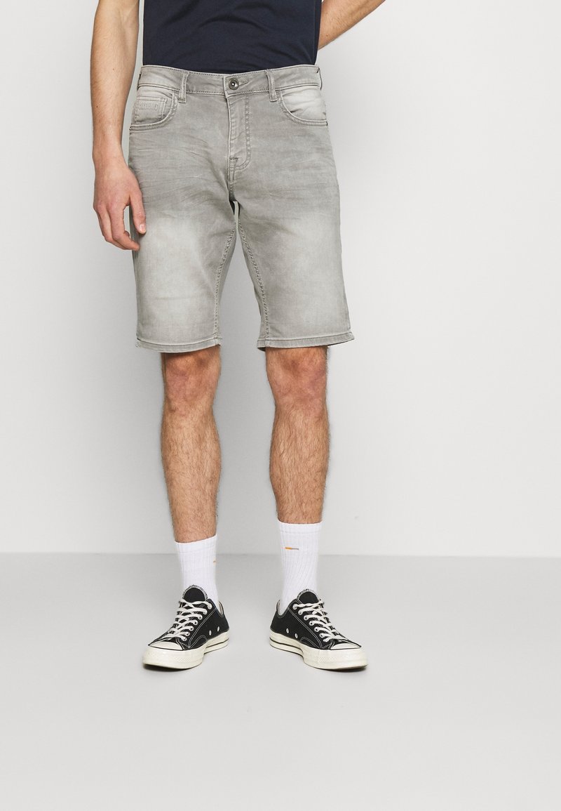 Man som bär urtvättade grå denimshorts, vita ankelsockor och svarta låga sneakers, stående mot en helt vit bakgrund.