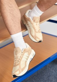Sneaker atletiche beige realizzate in rete e materiali sintetici, con tre righe bianche, una suola testurizzata e una linguetta imbottita con logo.