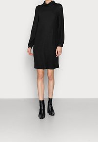 Černé dlouhé šaty s cowl krkem, vyrobené z měkké tkaniny; do poloviny kolen s volným střihem; doplněné černými kotníkovými botami na podpatku.